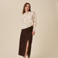 A woman modeling a cream knit sweater with a brown abstract pattern and a dark brown midi skirt with a front slit, paired with dark brown pointed-toe flats, against a solid beige studio background.