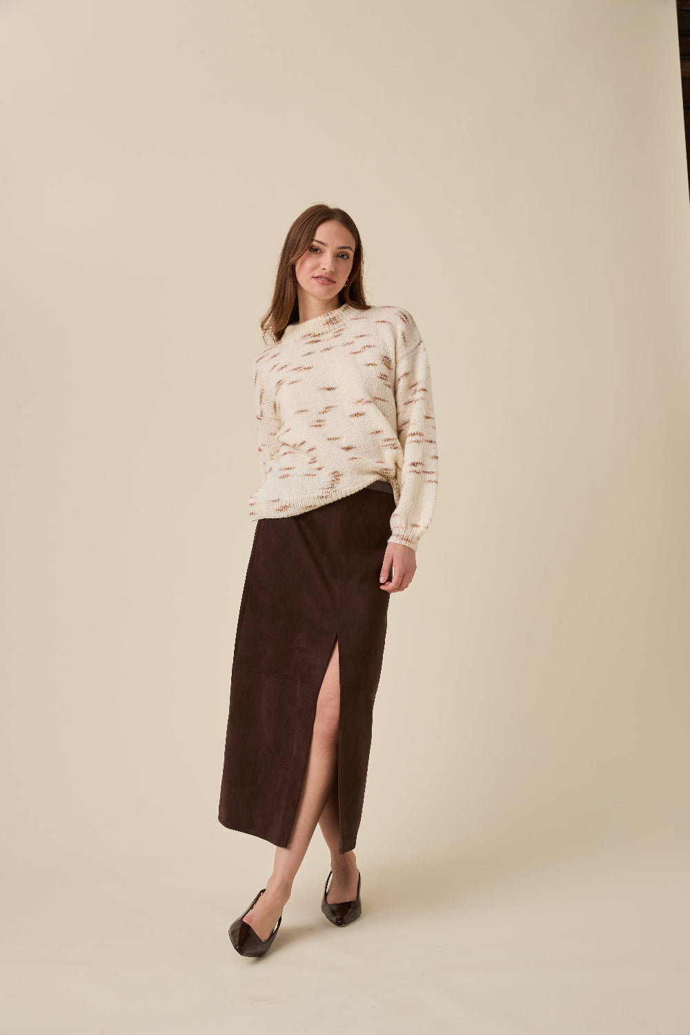 A woman modeling a cream knit sweater with a brown abstract pattern and a dark brown midi skirt with a front slit, paired with dark brown pointed-toe flats, against a solid beige studio background.
