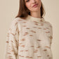A woman wearing a cream-colored knit sweater with brown and rust-colored abstract horizontal streaks, photographed against a solid beige background.