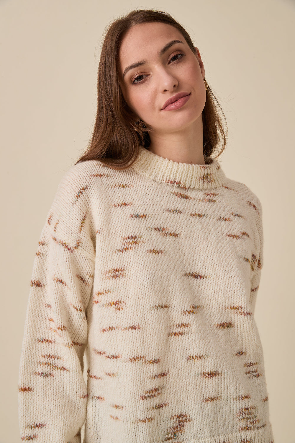 A woman wearing a cream-colored knit sweater with brown and rust-colored abstract horizontal streaks, photographed against a solid beige background.