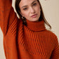 A woman wearing a chunky knit turtleneck sweater in a warm rust orange color, posed against a solid beige background.