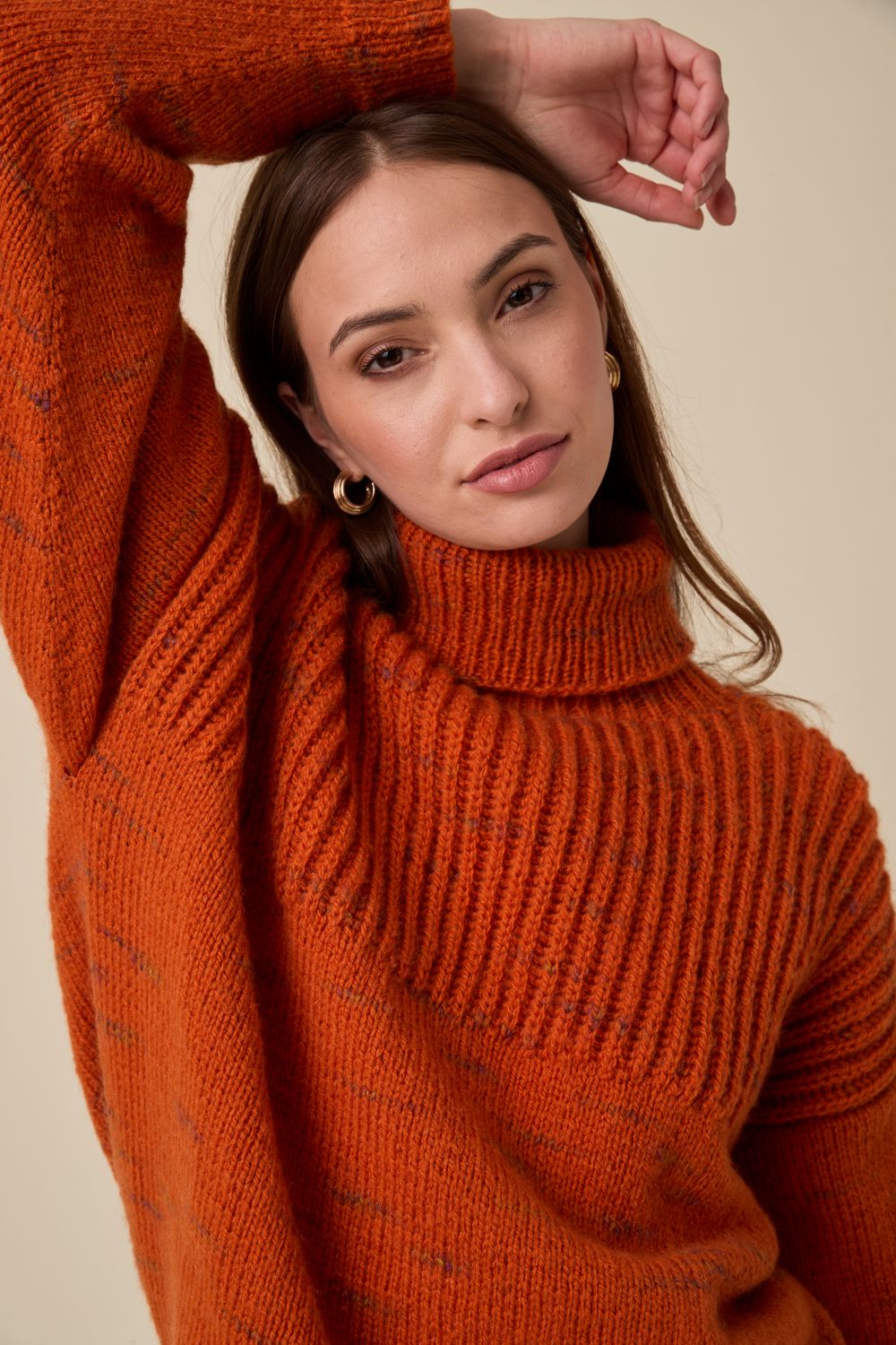 A woman wearing a chunky knit turtleneck sweater in a warm rust orange color, posed against a solid beige background.