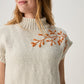 A woman wearing a cream-colored knit mock-neck sweater with orange embroidered leafy branches on the chest, photographed against a neutral light gray background.