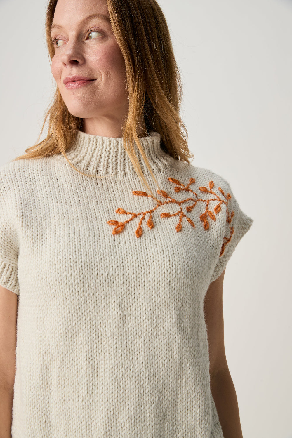 A woman wearing a cream-colored knit mock-neck sweater with orange embroidered leafy branches on the chest, photographed against a neutral light gray background.